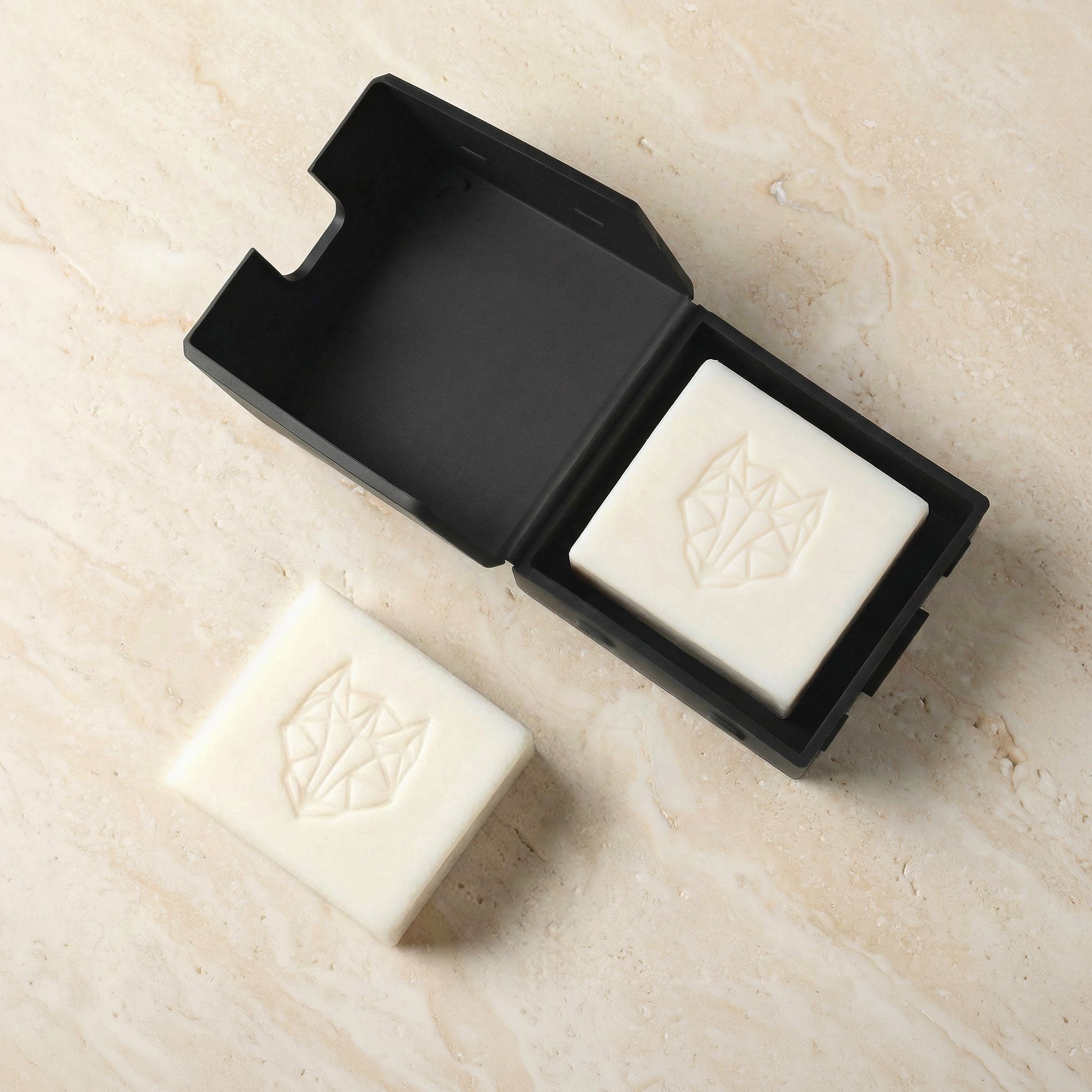 Two bars of soap with a black silicone travel case on a beige surface